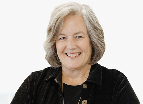 A smiling older woman with gray hair wearing a black jacket and a black top, standing against a plain white background.