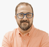 A smiling man with short brown hair, a beard, and blue glasses, wearing a light orange collared shirt, poses in front of a plain white background.