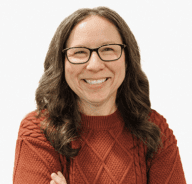 A smiling woman with long brown hair, wearing glasses and a rust-colored knit sweater, stands with her arms crossed against a plain white background.