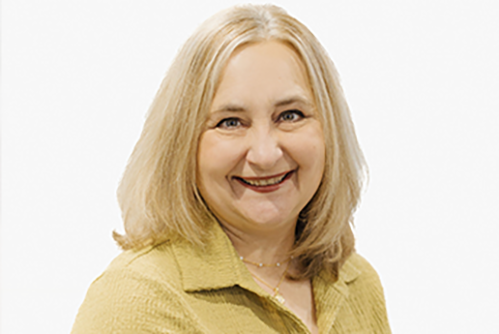 A smiling woman with straight, shoulder-length blonde hair wearing a light yellow collared top, posing against a plain white background.