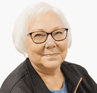 An older woman with short white hair, glasses, and a friendly smile is wearing a dark jacket over a blue top, posed in front of a plain white background.