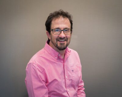 Jeff poses for a head shot in front of a grey background wearing a pink button-up shirt