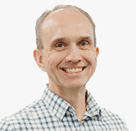 A man with short light hair, wearing a blue and white checkered button-up shirt, smiles at the camera against a plain white background.