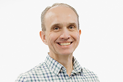 A man with short light hair, wearing a blue and white checkered button-up shirt, smiles at the camera against a plain white background.