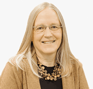 A smiling woman with long, straight blonde hair, wearing glasses, a brown cardigan, a black top, and a chunky wooden bead necklace, posed against a plain white background.