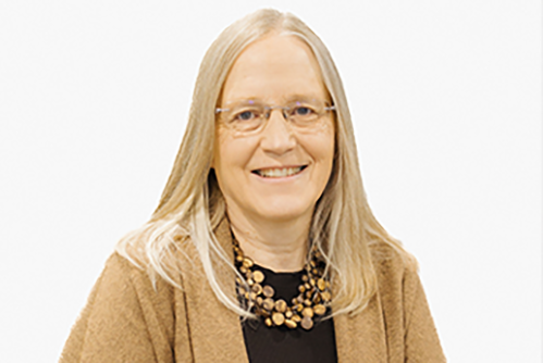 A smiling woman with long, straight blonde hair, wearing glasses, a brown cardigan, a black top, and a chunky wooden bead necklace, posed against a plain white background.