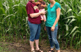 Grandma holds her infant granddaughter and stands next to her daughter-in-law at the edge of a field
