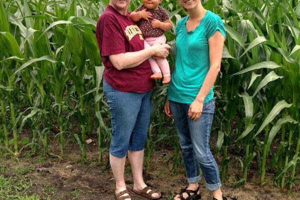 Grandma holds her infant granddaughter and stands next to her daughter-in-law at the edge of a field