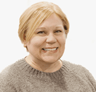 A woman with short blonde hair, wearing a light brown knit sweater, smiles at the camera against a plain white background.