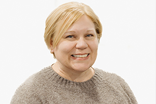 A woman with short blonde hair, wearing a light brown knit sweater, smiles at the camera against a plain white background.