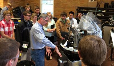 KCEO students gather in a semi-circle around a machine as Nova-Tech Engineering and MinnWest Technology Campus President Jim Sieben explains its function
