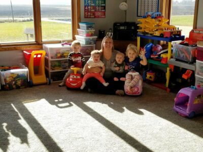 A group of kids is gathered around Michelle sitting on the carpet and surrounded by toys