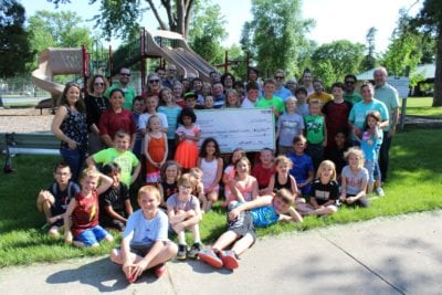 A large group of kids and adults gathers around a giant check at an outdoor park