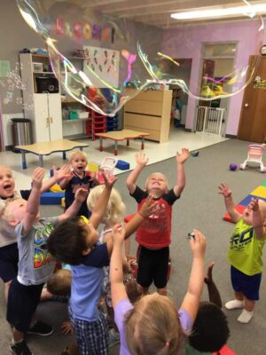 A group of young children hold their hands in the air trying to catch a giant bubble