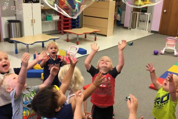 A group of young children hold their hands in the air trying to catch a giant bubble
