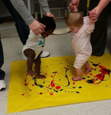Two babies walk on a large piece of yellow paper covered in paint