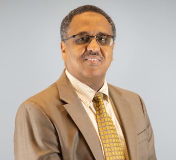 Zack is seated in front of a gray background and is wearing a brown suit coat with a bright yellow patterned tie.