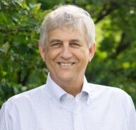 Tom stands outside in front of a forested background, wearing a lightly patterned button-up shirt.