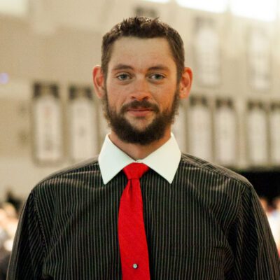 Matthew is pictured in a striped shirt and red tie in the middle of a gymnasium.