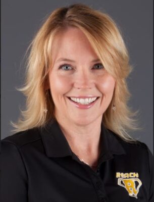 Rhoda sits for a formal head shot wearing a black polo with the REACH logo in yellow.