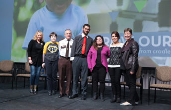 Seven people stand in a row on stage in front of a large projected image of a child at the Grow Our Own summit