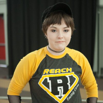 Teddy is pictured wearing a yellow and black REACH shirt in front of a black curtain backdrop.