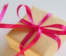 A small square gift is wrapped in brown kraft paper and wound in a bright pink ribbon tied with a bow.