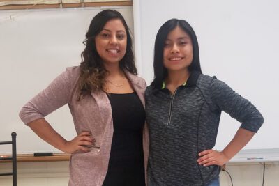 Perla and Jessica stand next to one another in front of a whiteboard in a classroom, each with a hand on her hip