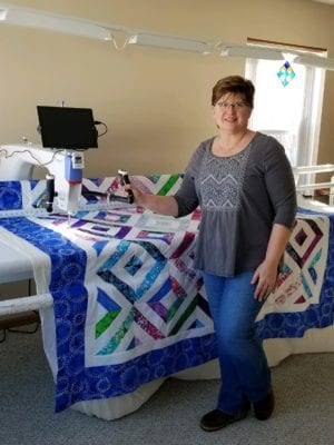 Rhonda stands next to the long arm quilting machine with a colorfully patterned quilt lying on it