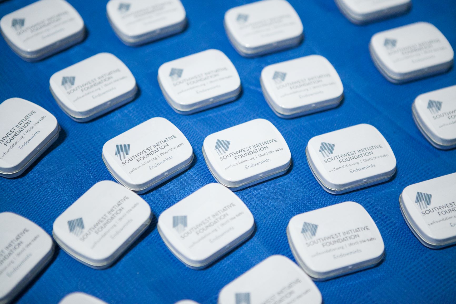 Small tins of mints with the SWIF logo emblazoned on the front are arranged in a pattern on a blue tablecloth
