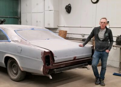 Rick is inside a shop, leaning with one hand on the sanded-down body of a classic car.