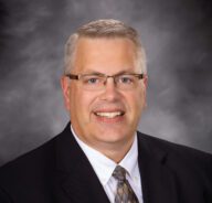 Dan poses for a formal head shot in front of a mottle gray backdrop