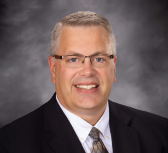 Dan poses for a formal head shot in front of a mottle gray backdrop