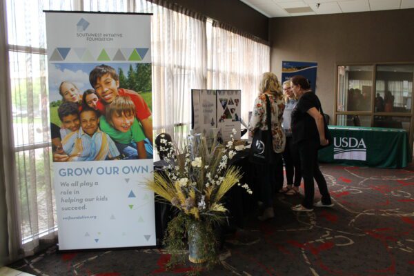 A banner with the words "Grow Our Own" at the top and a group of kids in brightly colored shirts stands next to people talking in a small group