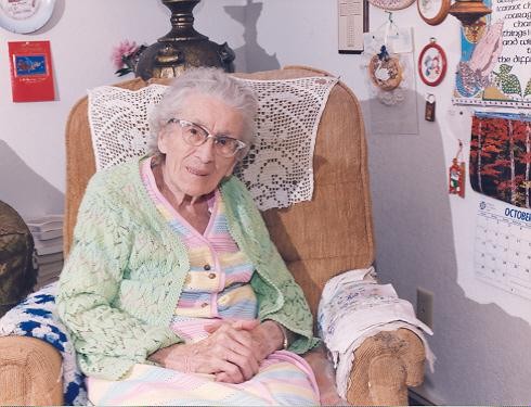 Alma sits in a tan arm chair with her hands crossed and resting on a quilt covering her lap