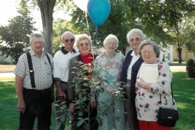 A group of men and women stand around a sapling with balloons tied to it