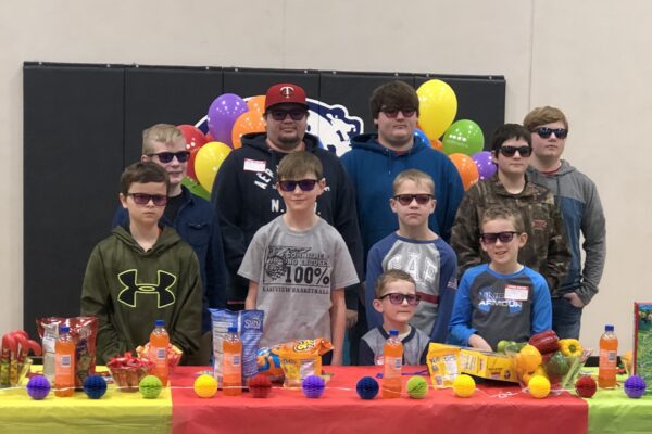 A group of students wearing glasses stand in front of a table covered in colorful items