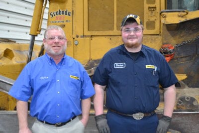 Chris and Parker stand next to each other in front of a large piece of equipment in the shop