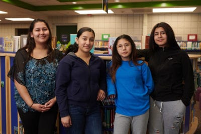 Four sisters stand shoulder-to-shoulder in front of bookshelves in a school library