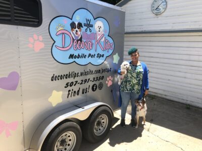 Kathryn stands next to her branded mobile pet spa with a small white dog tucked under her arm as she holds the leash of a white and brown dog standing next to her
