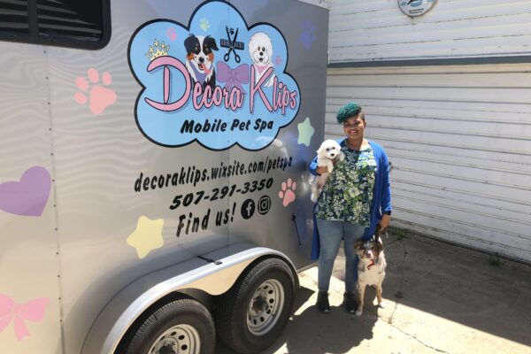 Kathryn stands next to her branded mobile pet spa with a small white dog tucked under her arm as she holds the leash of a white and brown dog standing next to her