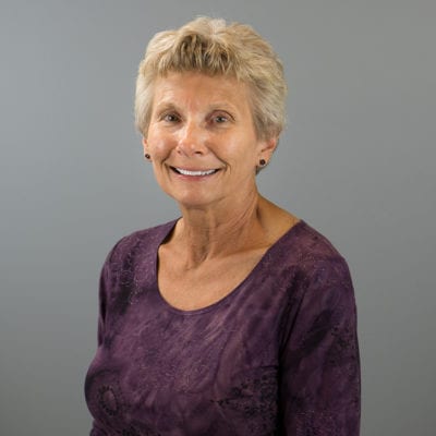 Marsha is wearing a purple shirt, posing for a formal head shot in front of a grey background