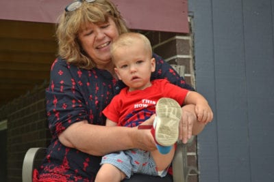 A woman sitting outside a blue house holds a small boy on her lap while she pulls on one of his shoes.