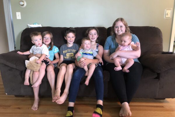 Makayla holds an infant and sits on a brown sofa with a group of kids