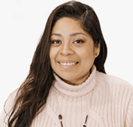A woman with long dark hair smiles at the camera. She is wearing a light pink ribbed turtleneck sweater and a necklace, with a plain white background behind her.