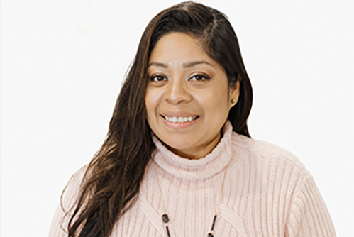 A woman with long dark hair smiles at the camera. She is wearing a light pink ribbed turtleneck sweater and a necklace, with a plain white background behind her.