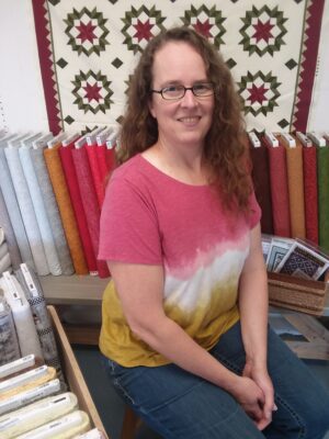 Lori sits in front of bolts of quilt fabric with a starburst wall quilt hanging behind her