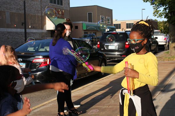 A young black girl in a yellow sweater creates giant bubbles on a sidwalk that another girl is chasing