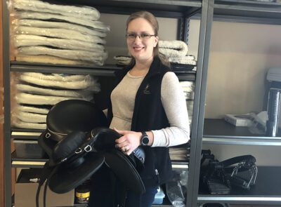 Dana holds a black saddle and stands in front of storage racking stacked with saddle inventory