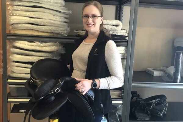 Dana holds a black saddle and stands in front of storage racking stacked with saddle inventory